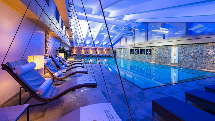 Der Raum zeigt einen modernen Innenpool mit blauem Licht und entspannenden Liegen. Die Atmosphäre ist ruhig und einladend, ideal für Entspannung und Wellness. Steinwände und Pflanzen tragen zum stilvollen Ambiente bei.